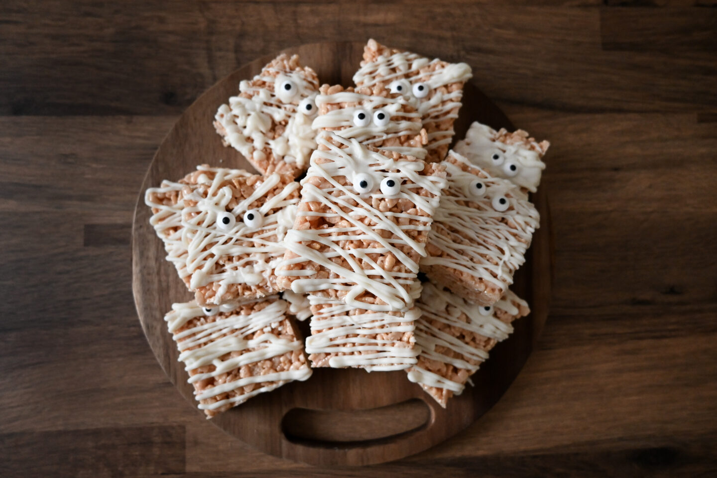 Mummy Rice Krispy Treats with edible eyeballs and white icing that looks like bandgaes on a round, wooden cutting board on a wooden table