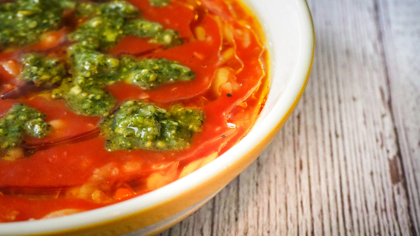 Close up of the Bowl of tomato, chickpea & orzo soup with Pistachio Pesto on a rustic white wooden background. The bowl is yellow and white striped cornishware