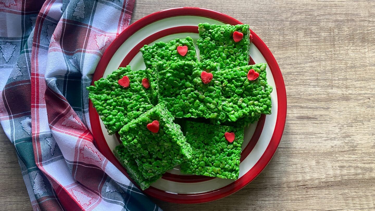 Grinch-themed Rice Krispy Treats that are green with little red hearts on a plate with a Christmas tree tea towel