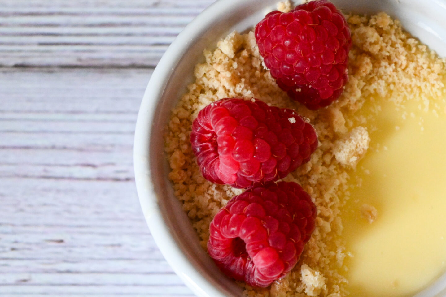Small white ramekin with lemon posset topped with shortbread crumb and fresh raspberries on a white background.