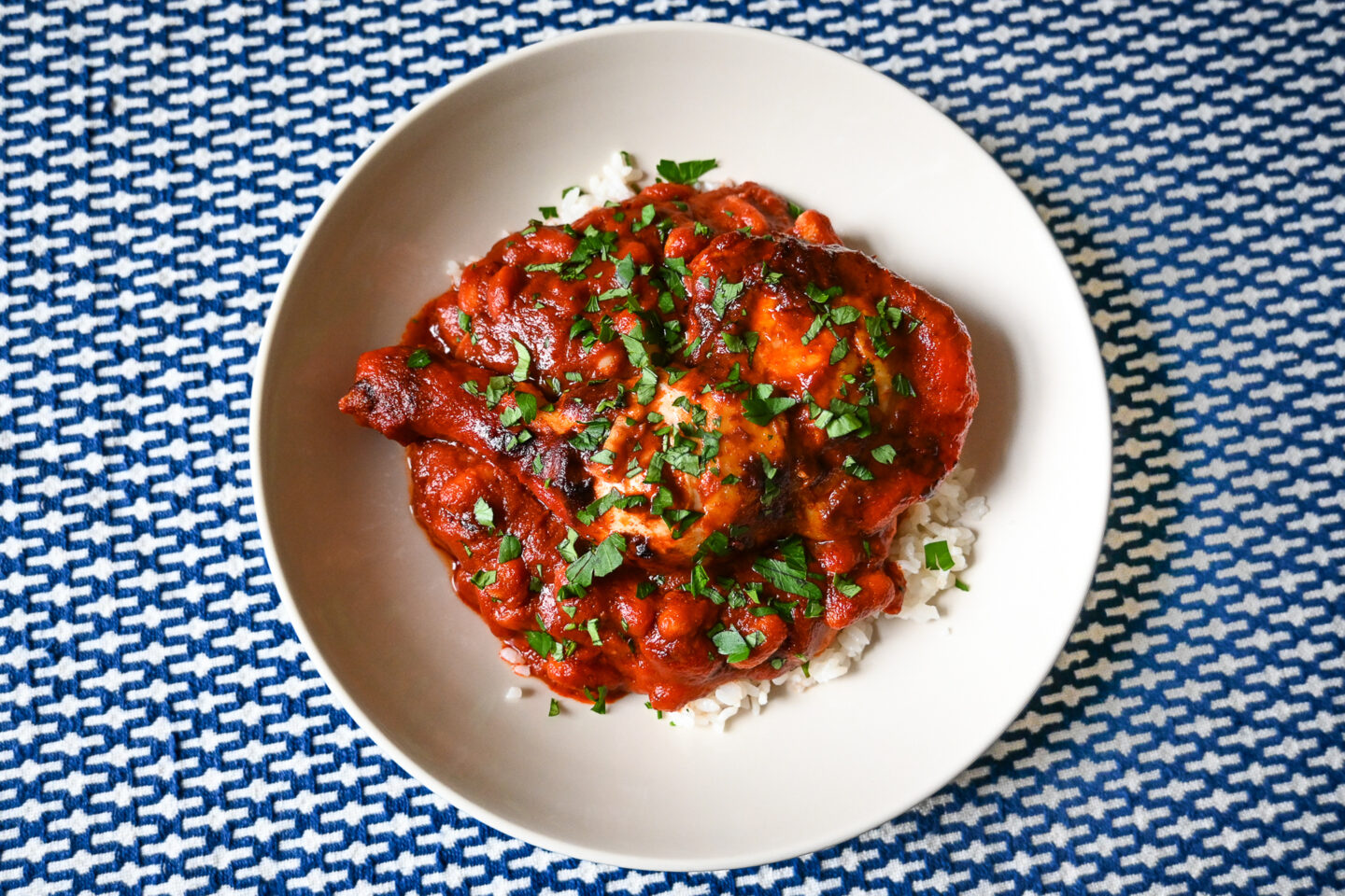 White bowl with red paprika chicken & cannellini Beans on a bed of white rice against a blue & white woven tablecloth