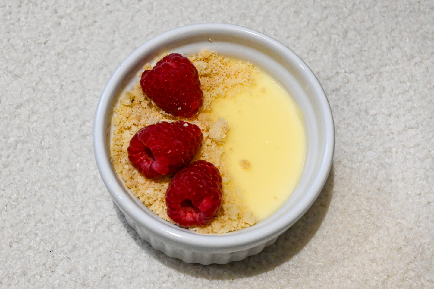 Lemon posset in a white ramekin with shortbread crumb and 3 raspberries on top