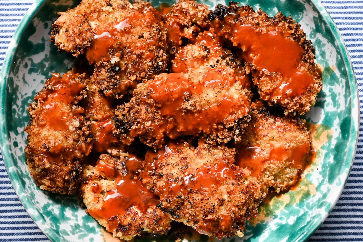 Crispy fried chicken tenders drizzled with buffalo sauce in a green and white speckled bowl on a blue and white striped background