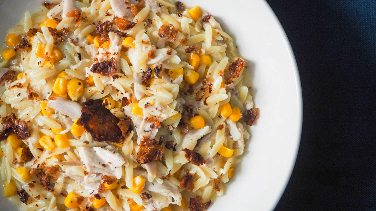 Chicken & Sweetcorn Orzo with Crispy Chicken Skin Sprinkled on top in a white pasta bowl on a dark background