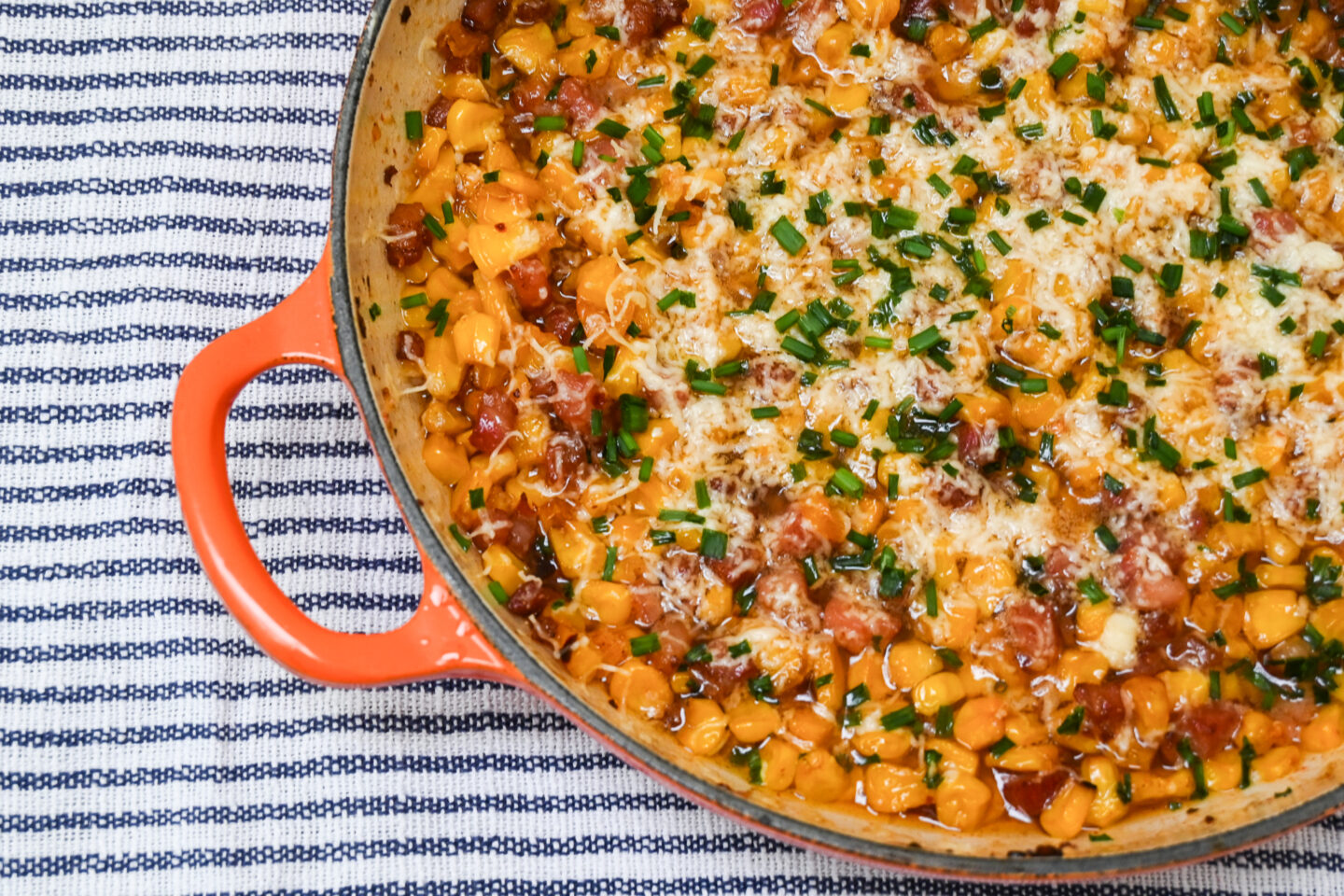 Orange Le Creuset Casserole with Corn & Pancetta topped with grated cheddar cheese and chives on a striped grey & white background