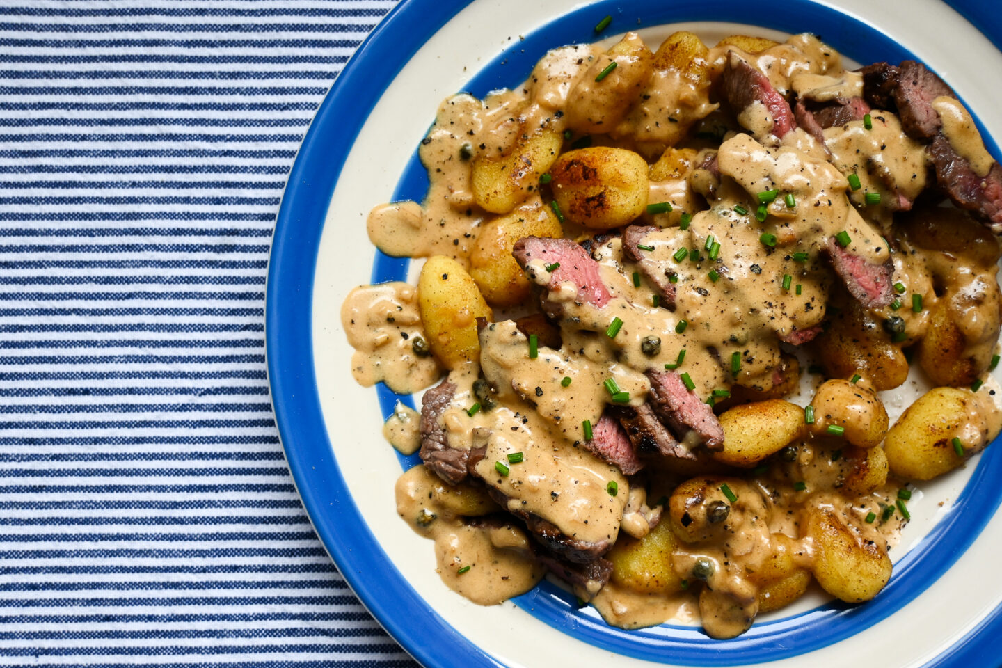 Fried Gnocchi with sliced medium-rare rump steak across the top drizzled with peppercorn sauce & sprinkled with chopped chives on a blue & white striped plate against a blue & white striped background.