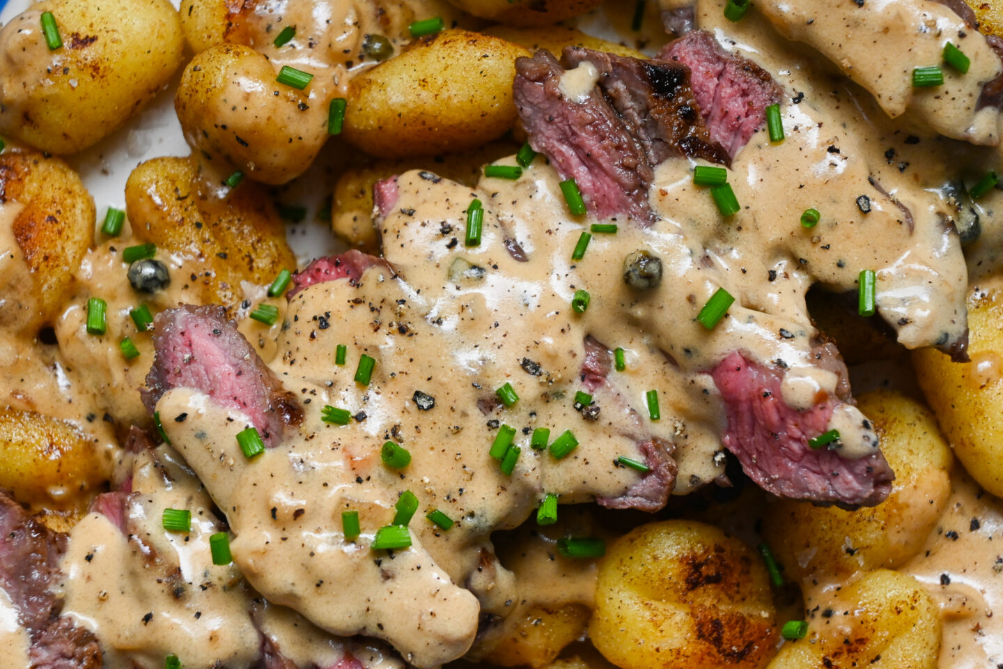 Close-up of medium-rare sliced rump steak over fried gnocchi with peppercorn sauce and chopped chives across the top.