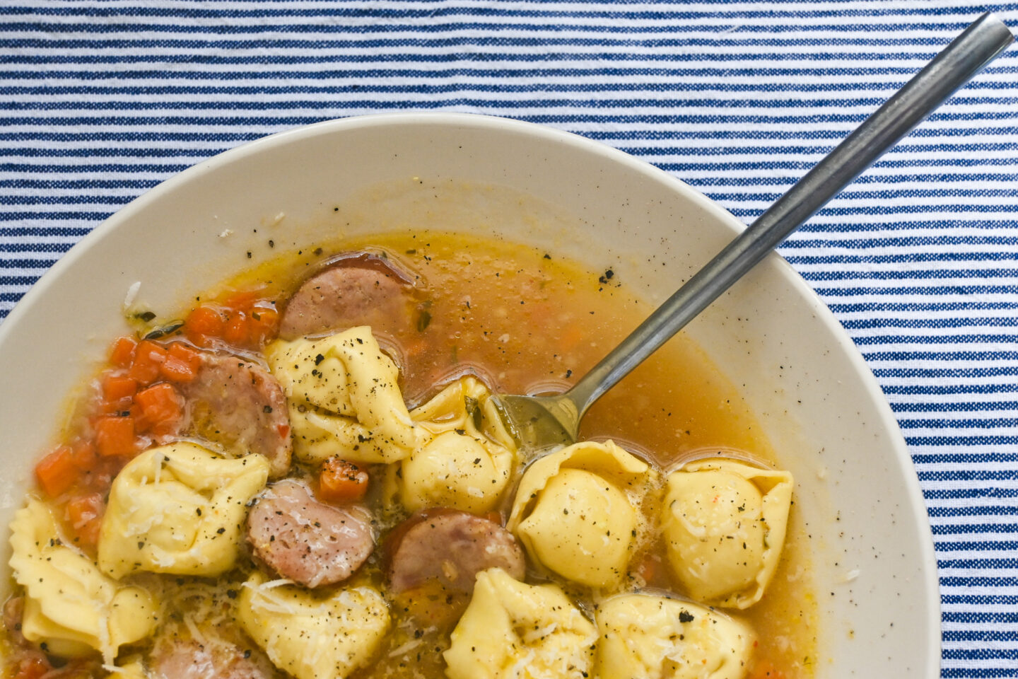 Tortelloni & Polish Sausage Soup With Diced Carrots in a white bowl on a blue and white striped background