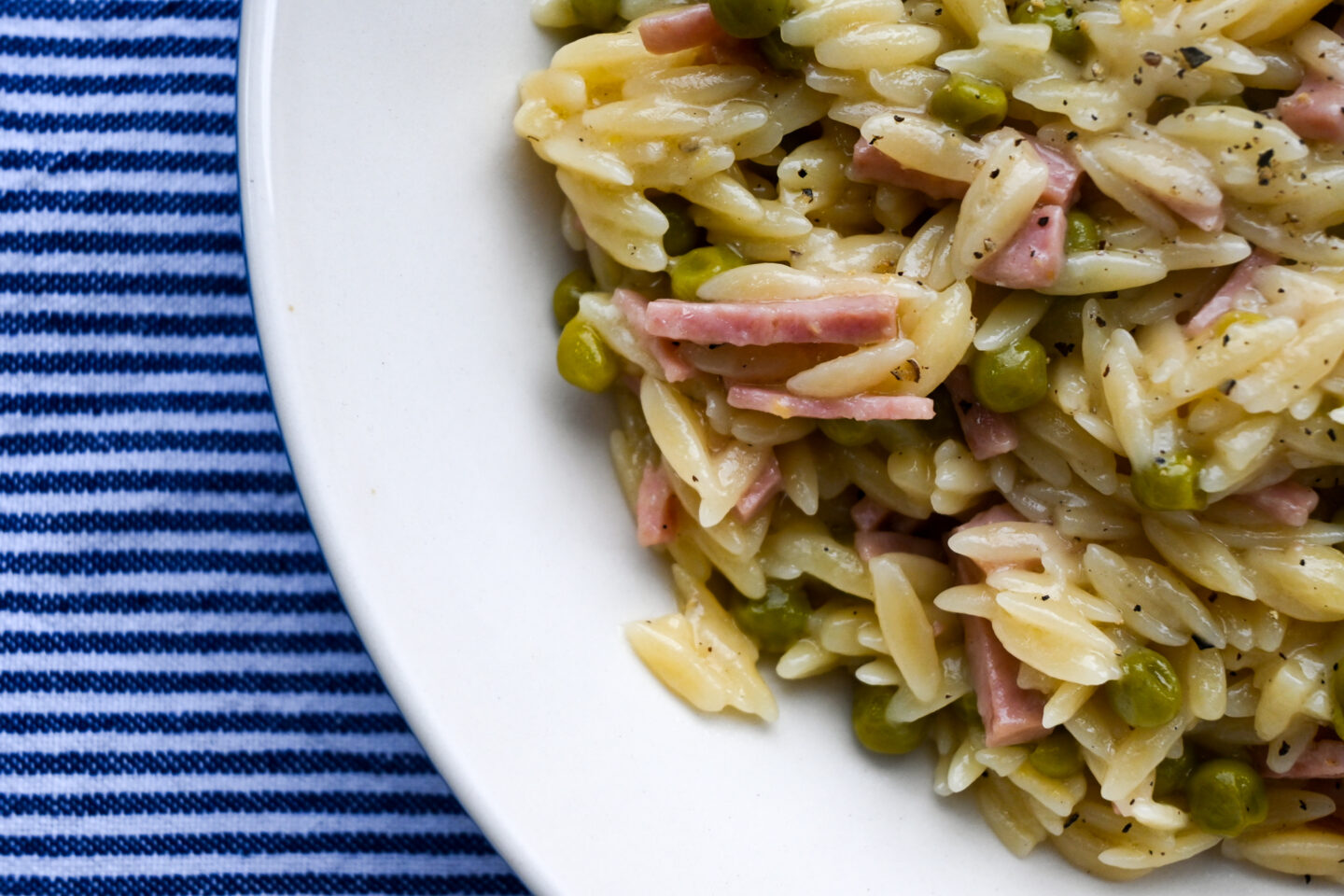 Corner of a plate of orzo, ham cubes & peas on a blue and white striped background