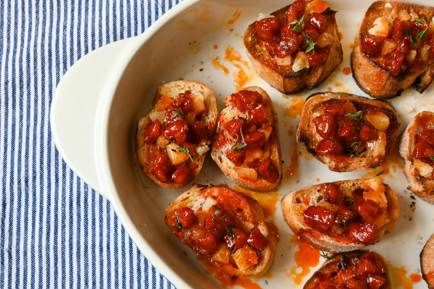 Honey Glazed Chorizo with Manchego Cheese on small slices of toasted baguette on a white dish with a blue & white striped background