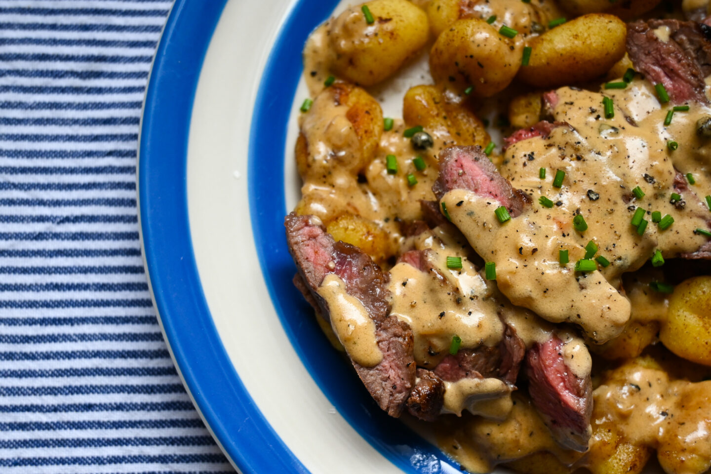 Close-up of fried Gnocchi with sliced medium-rare rump steak across the top drizzled with peppercorn sauce & sprinkled with chopped chives on a blue & white striped plate against a blue & white striped background.