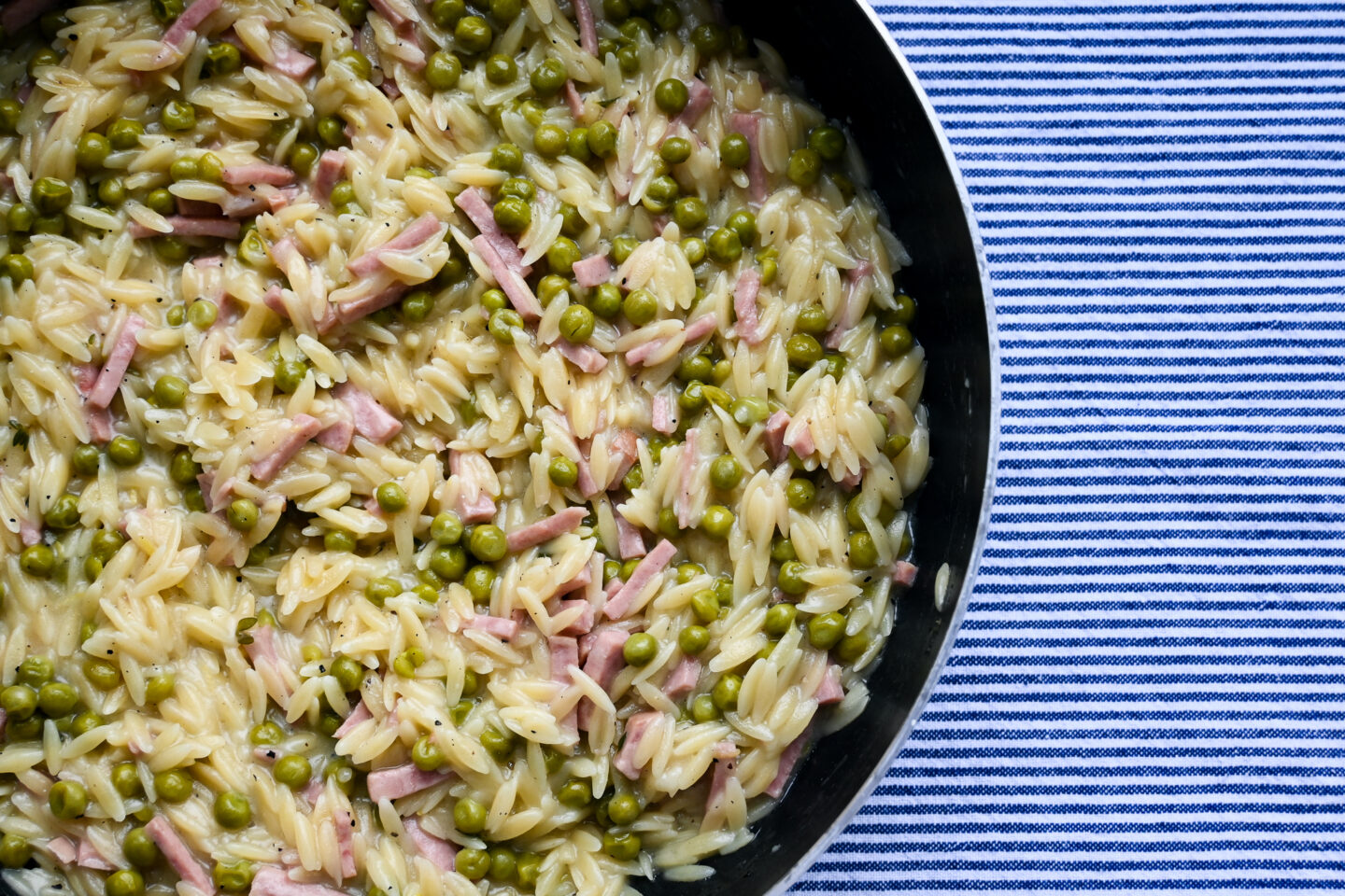 Skillet with orzo, ham cubes & peas on a blue and white striped background