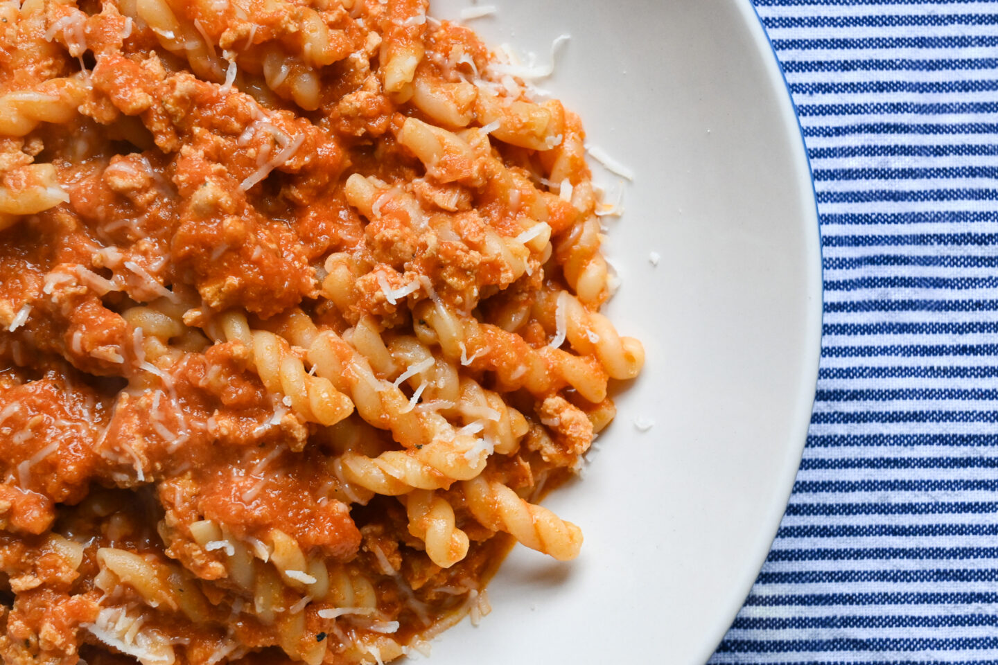 Gemelli Pasta With Chicken Bolognese Sauce sprinkled with Parmesan cheese over top on a white plate with a blue & white striped background