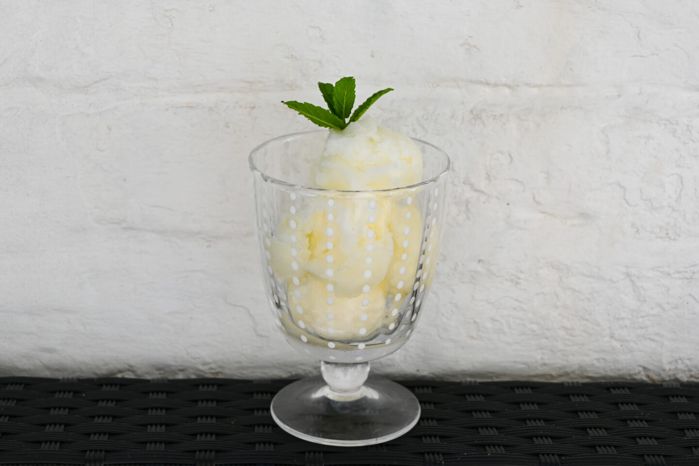 Scoops of fizzy lemon sorbet in a parfait glass with a sprig of mint on top against a white wall background