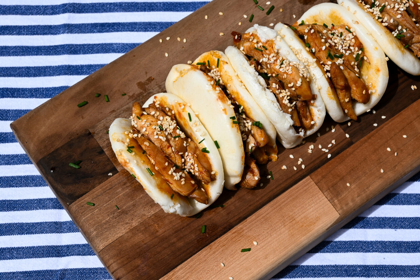 Honey, Soy & Harissa Chicken Bao Buns on a dark wooden cutting board sprinkled with chives & sesame seeds on a blue & white striped background