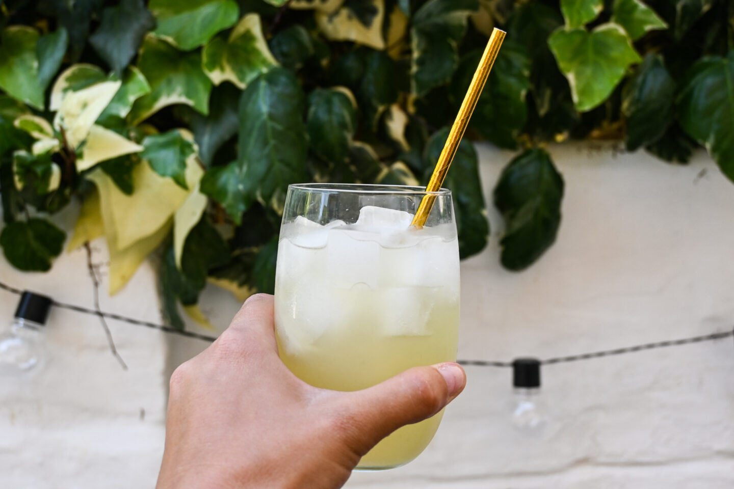 Hand holding a clear glass of lemonade with ice and a gold straw against a white garden wall with ivy and outdoor lights strand