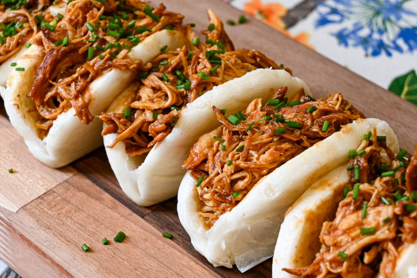 Bao buns stuffed with pulled bbq chicken and sprinkled with chopped chives on a wooden serving board sitting on a floral table cloth