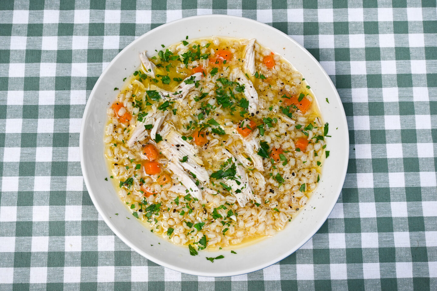 Chicken & barley soup with carrots & parsley in a wide, white bowl on a sage green & white gingham background