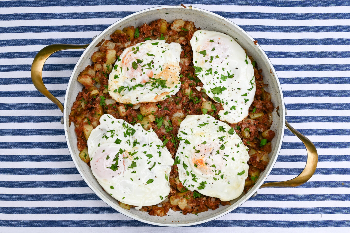 Corned Beef Hash topped with Fried Eggs and Sprinkled chopped parsley in a skillet on a blue & white striped background