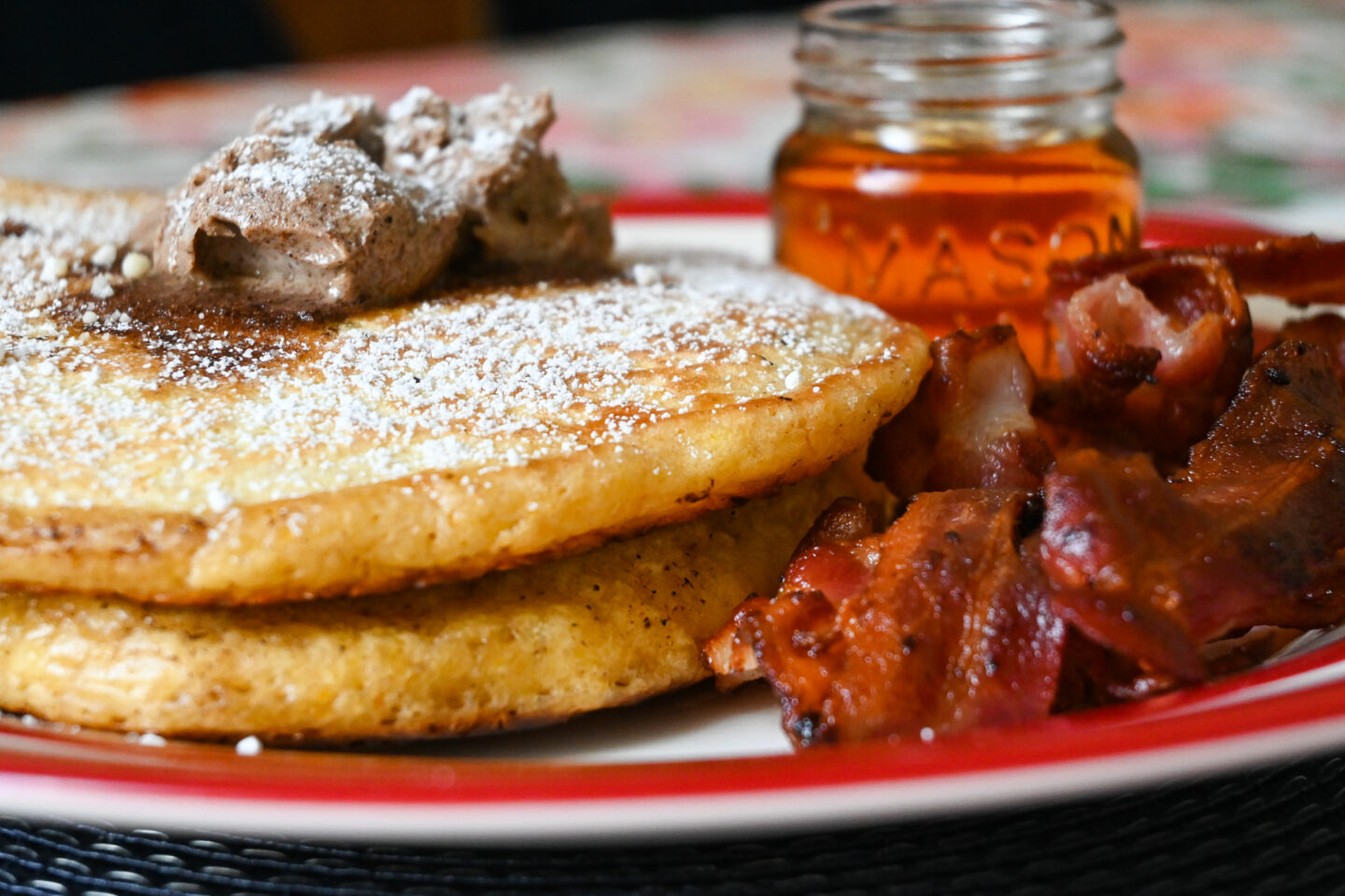 Closeup of fluffy orange pancakes with cinnamon butter on top and crispy streaky bacon & maple syrup in a small Mason Jar on a red and white plate