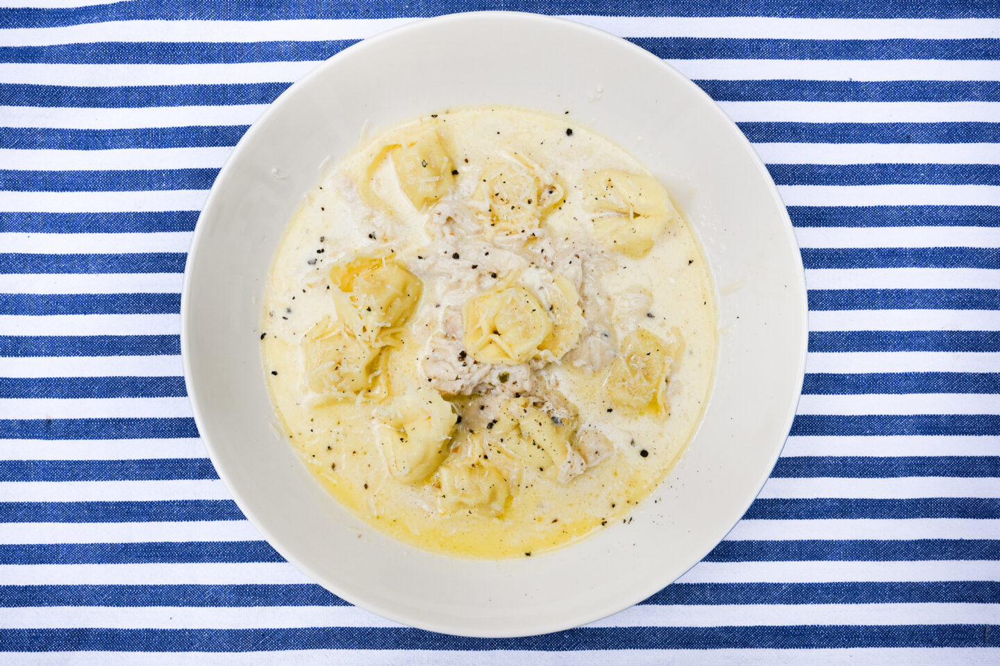 Bowl of creamy soup with pulled chicken and cheese tortelloni in a bowl on a blue & white striped background