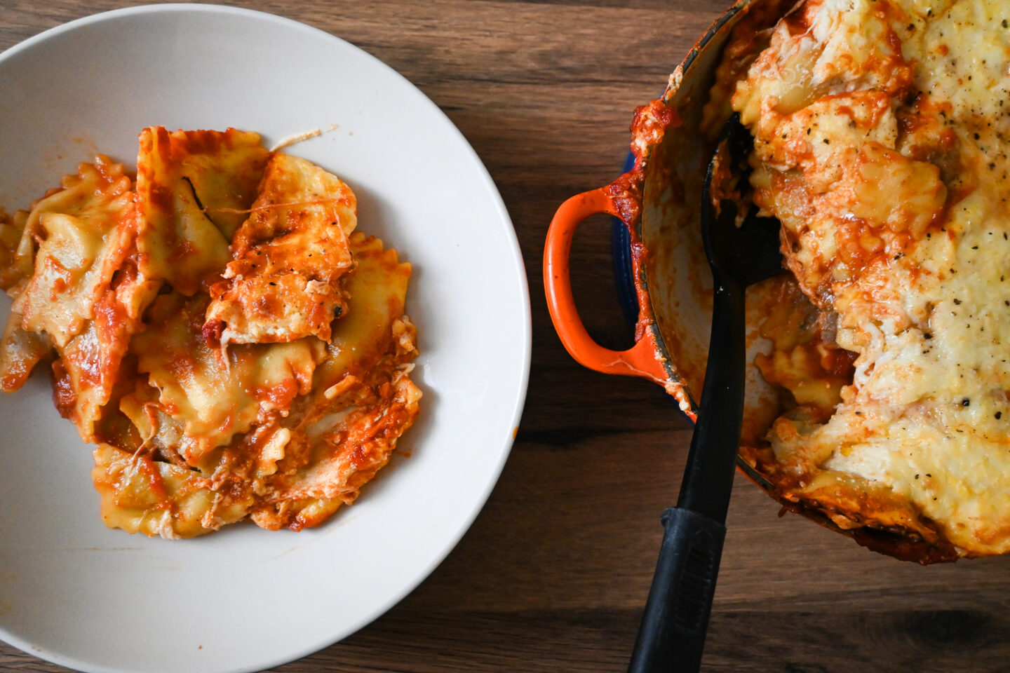 Cheesy Baked Ravioli in an orange casserole and a serving dished out into a white bowl on a wooden table