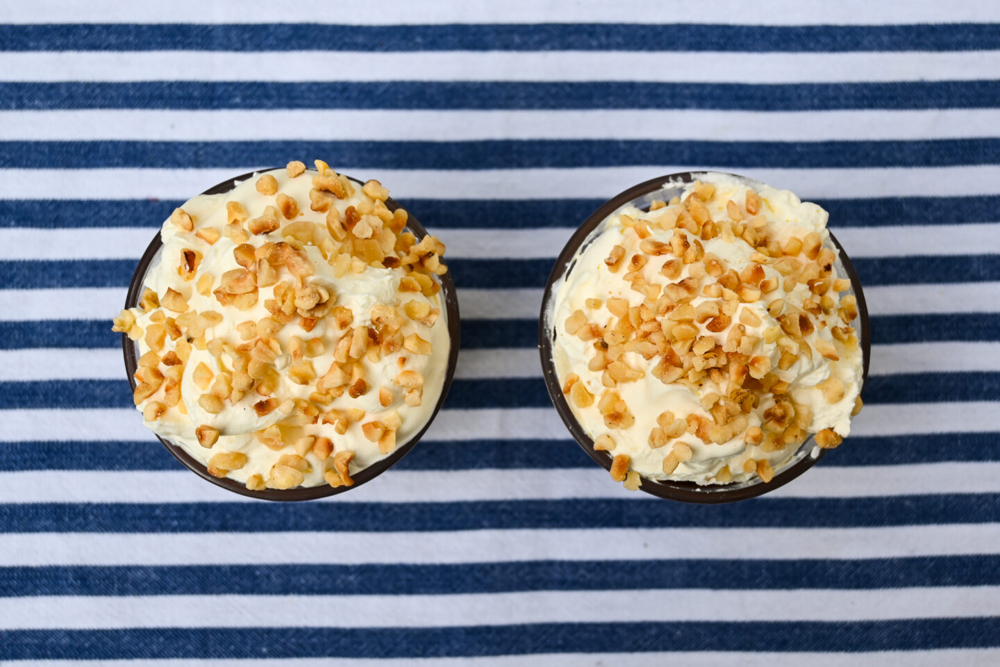 Top view of pudding filled glasses with whipped cream and nuts on top against a blue & white striped background