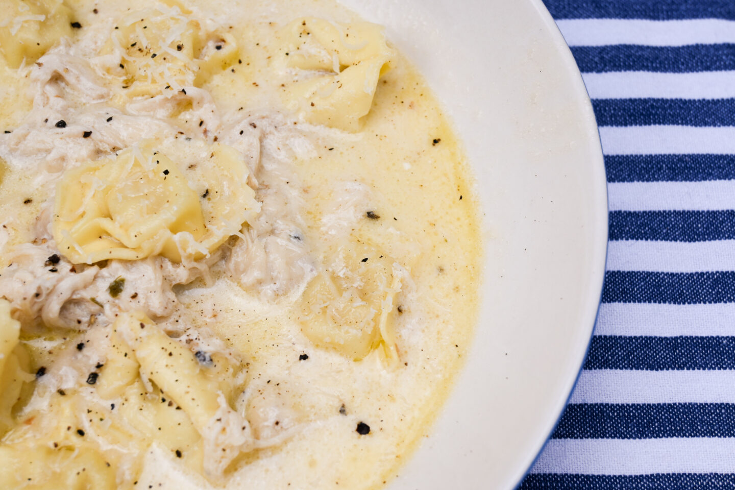 Bowl of creamy soup with  pulled chicken and cheese tortelloni in a bowl on a blue & white striped background closeup