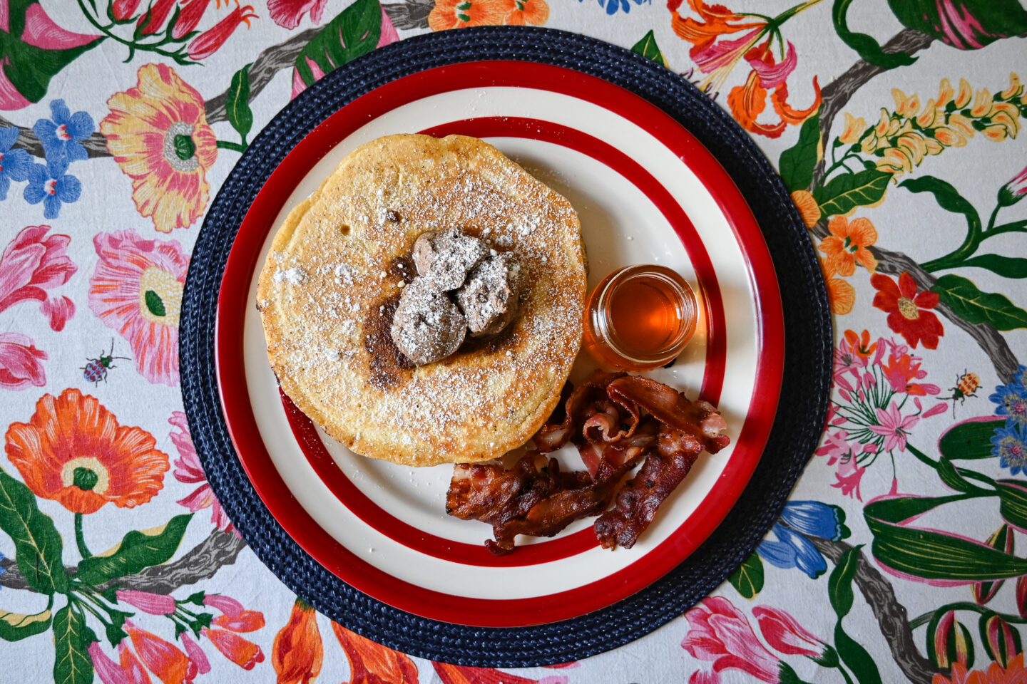 Shot from above of fluffy orange pancakes with cinnamon butter on top and crispy streaky bacon & maple syrup in a small Mason Jar on a red and white plate with a navy round placemat on a floral table cloth