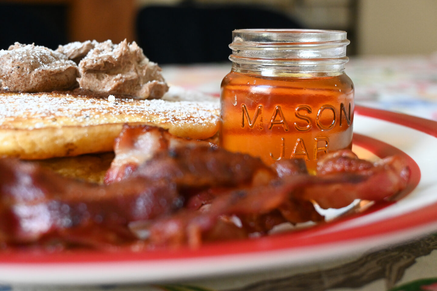 Closeup of fluffy pancakes, crispy streaky bacon & maple syrup in a small mason jar on a red & white plate