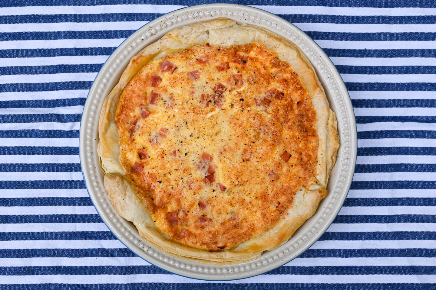 Top shot of whole puff pastry ham & cheese quiche in a le creuset stonewear pie plate on a blue and white background