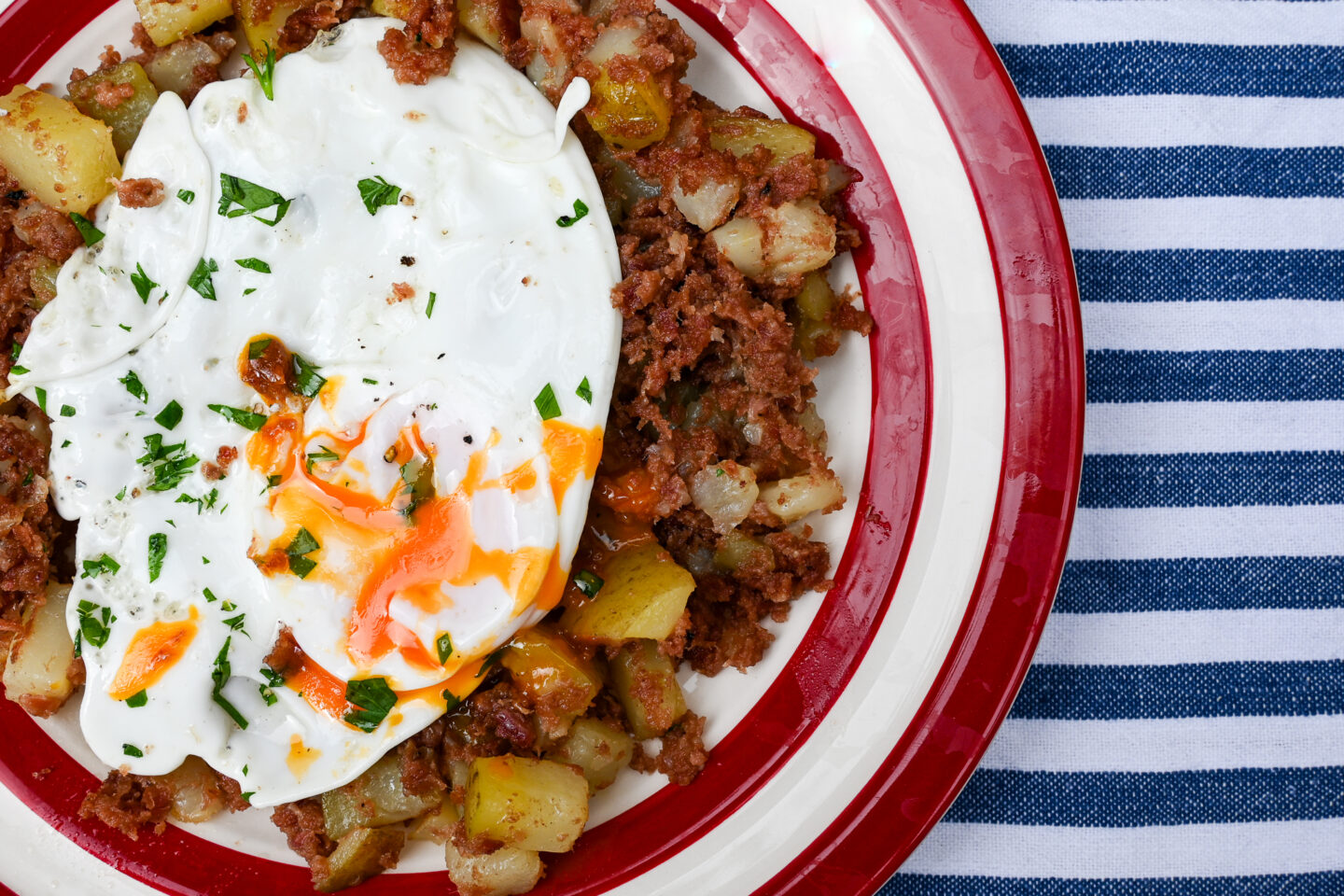 Corned beef hash with a runny fried egg on a red & white striped plate on a blue & white striped background