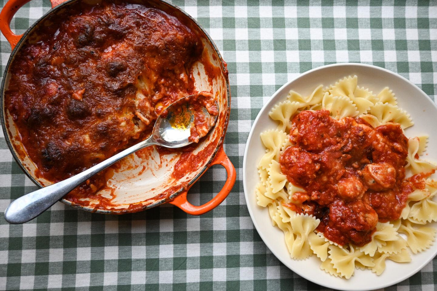 Oven Baked Cheesy Chicken Meatballs in an Orange Le Creuset Casserole with a silver serving spoon next to a plate of bowtie pasta with a serving of the meatballs on top.