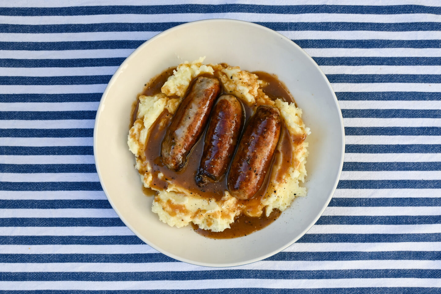 3 fat sausages on a bed of creamy mashed potatoes with a dark gravy poured over in a white plate on a blue & white Striped background