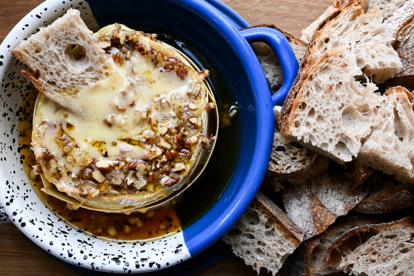 Wedge of bread dipped into baked camembert with maple & pecans on top in a blue dish with more bread on the side