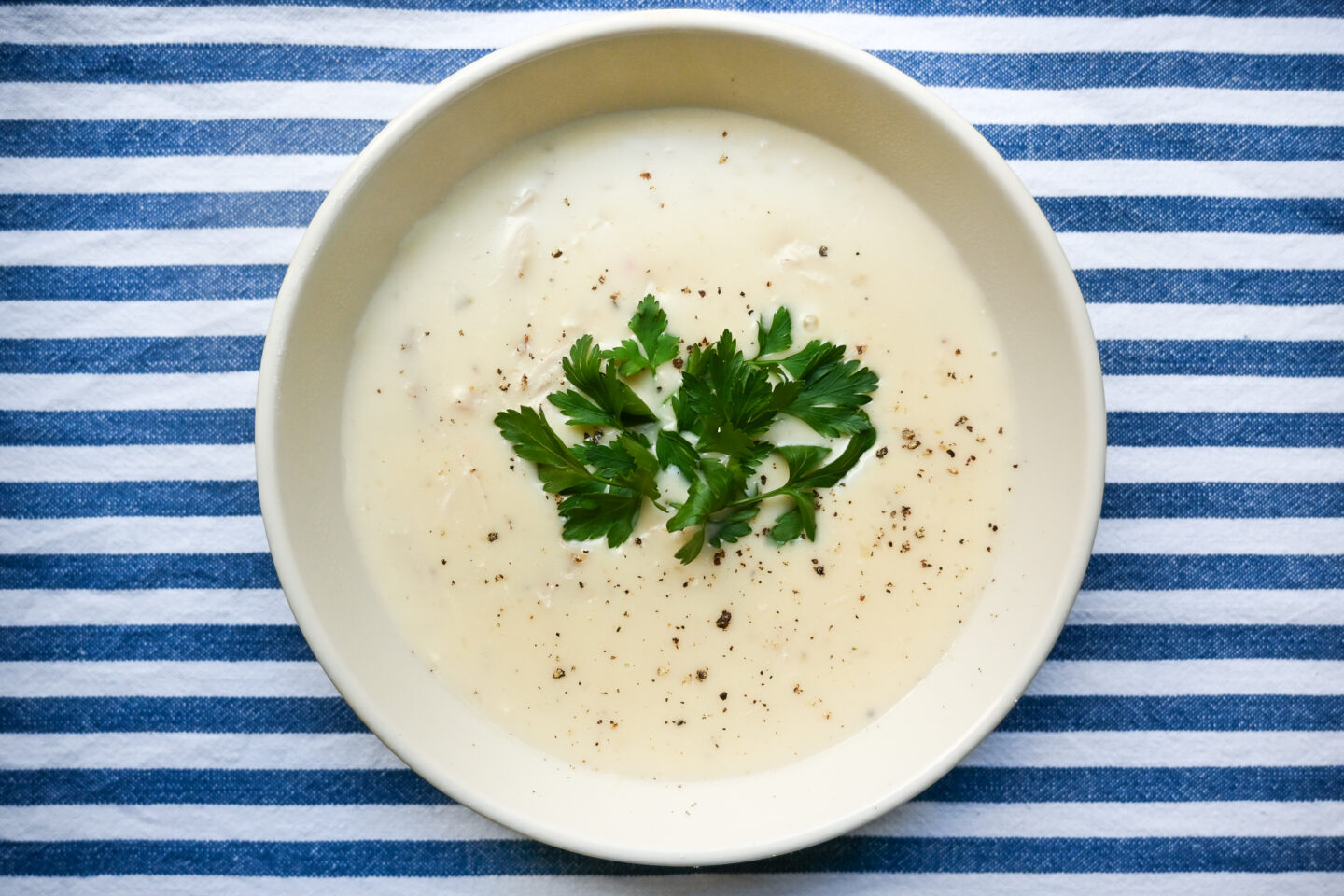 Bowl of cream of chicken soup with cracked black pepper and fresh parsley on top sitting on a blue & white striped background