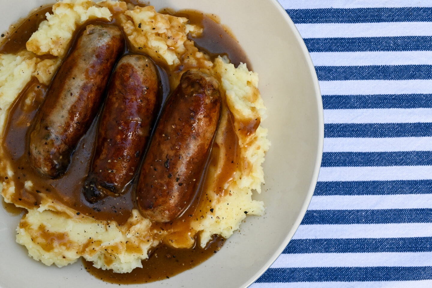 Side shot of sausages & mash with gravy on a white plate with a blue & white striped background