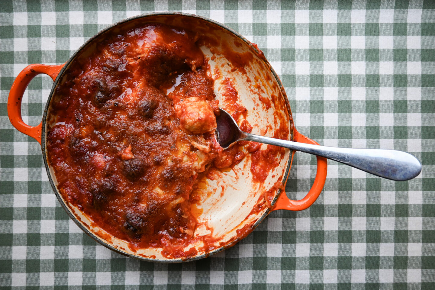 Orange Le Creuset Cast Iron shallow Casserole with Oven Baked Cheesy Chicken Meatballs and a Silver Serving Spoon on a green & white gingham background