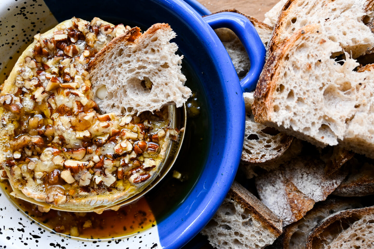 Wedge of bread dipped into baked camembert with maple & pecans on top in a blue dish with more bread on the side