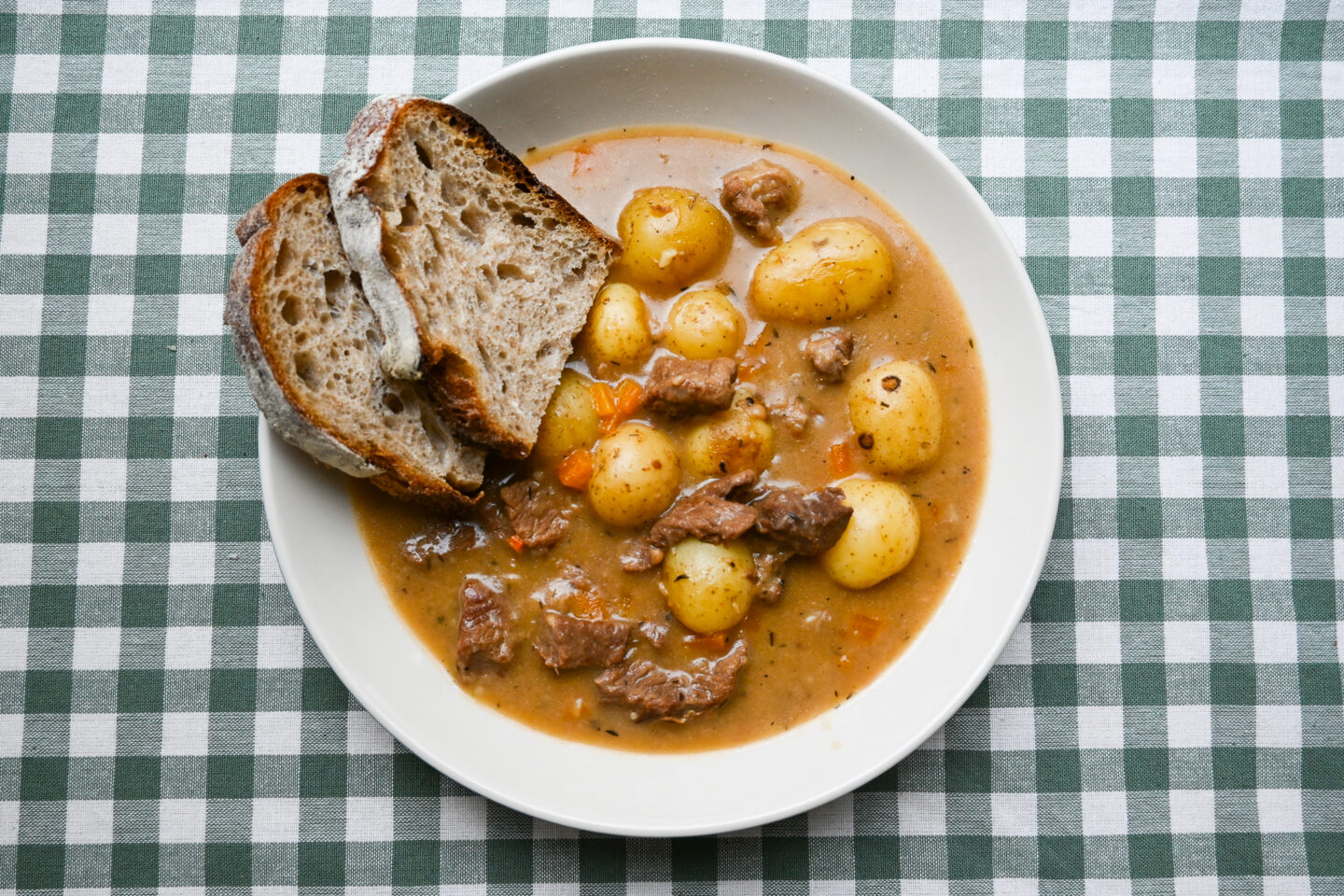 White wine & dijon beef stew with carrots & potatoes served in a white wide bowl with two slices of sourdough bread on a green & white gingham background