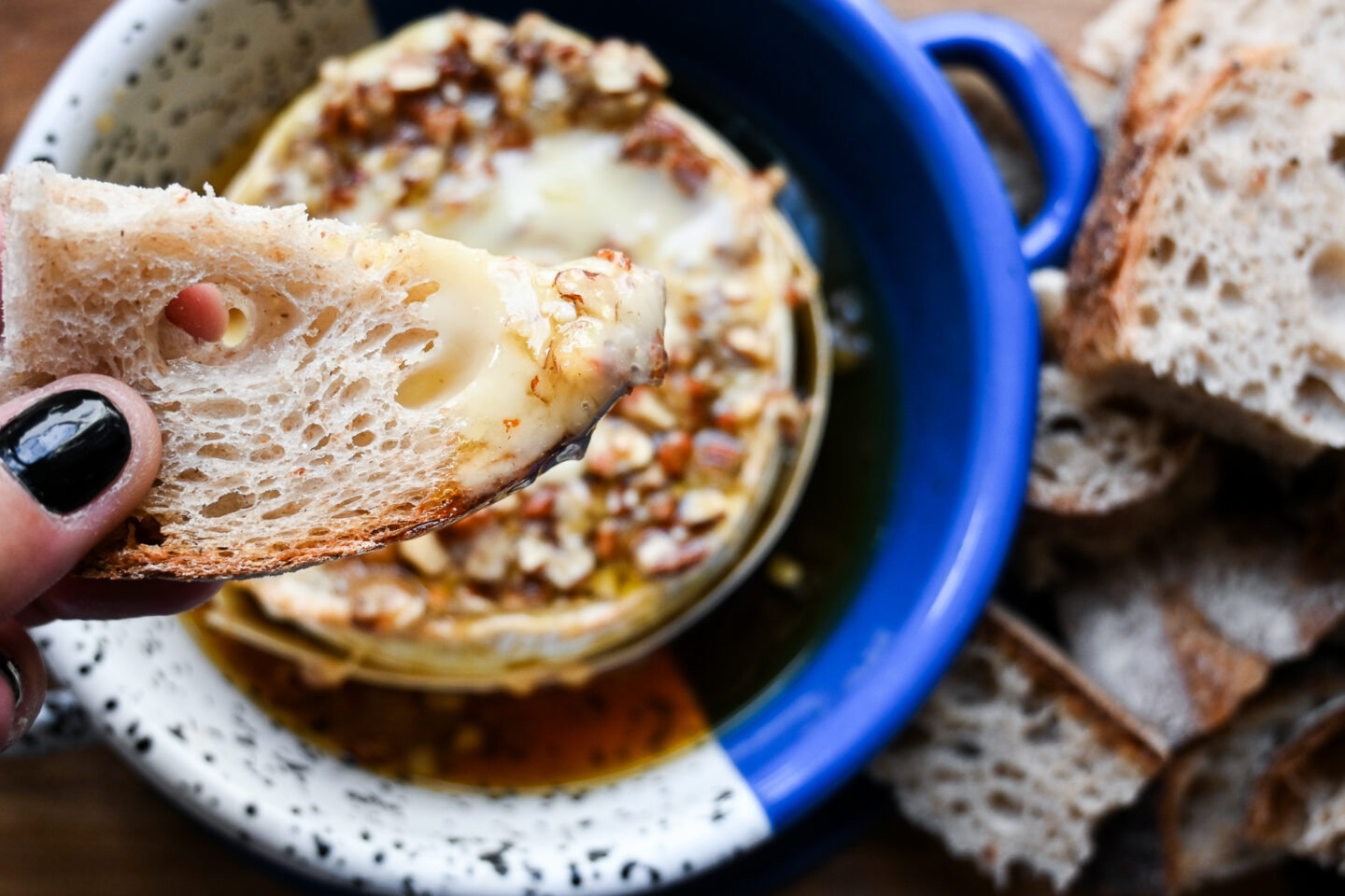 Wedge of bread dipped into baked camembert with maple & pecans on top in a blue dish with more bread on the side