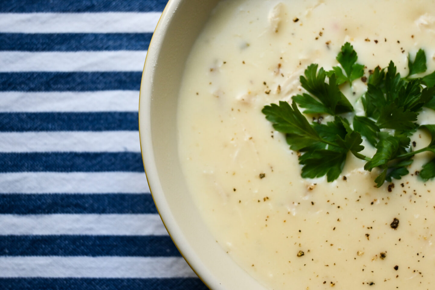 Side shot of cream of chicken soup garnished with black pepper & fresh parsley on top of a blue & white striped background