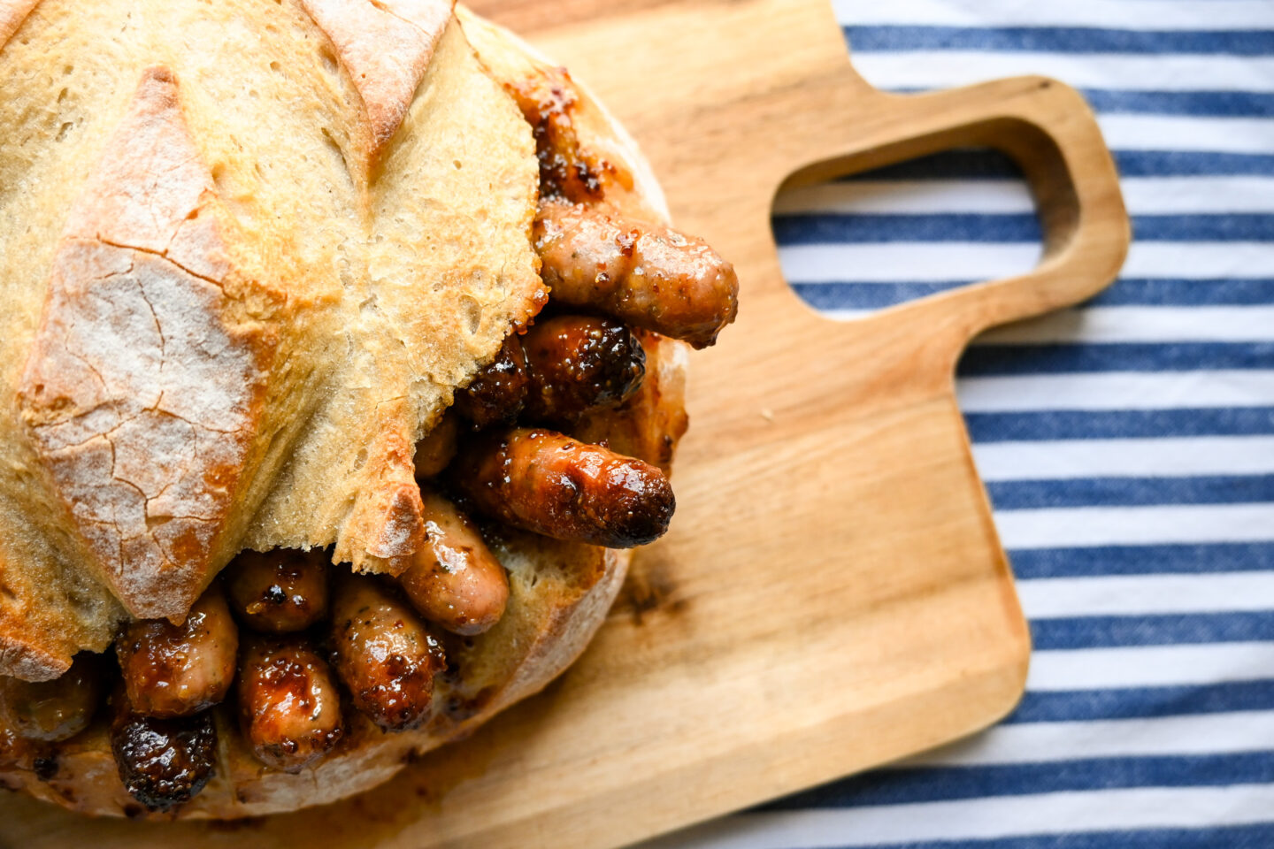 Honey mustard chipolatas peeking out of a bread bowl on a cutting board with a striped blue & white background