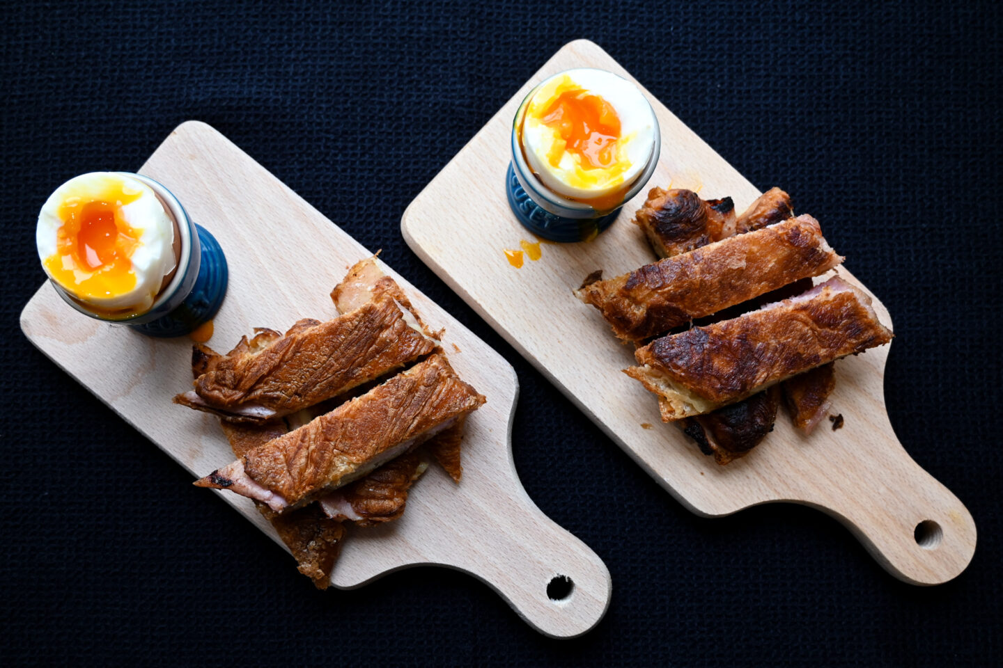 Soft Boiled Eggs in blue Le Creuset Egg Cups With Ham & Cheese Croissant Dippers on small handled cutting boards with a black background