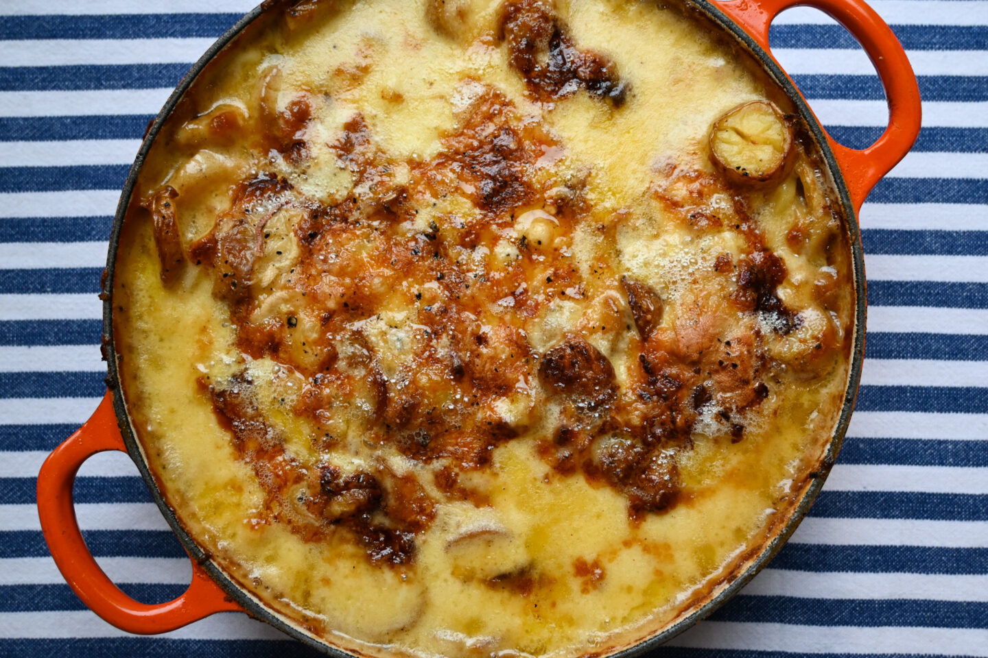 Tartiflette in an orange shallow Le Creuset Casserole on a blue and white striped background