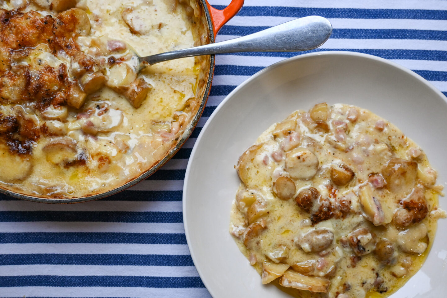 Serving of Tartiflette in a shallow bowl next to the orange casserole containing Tartiflette and a serving spoon