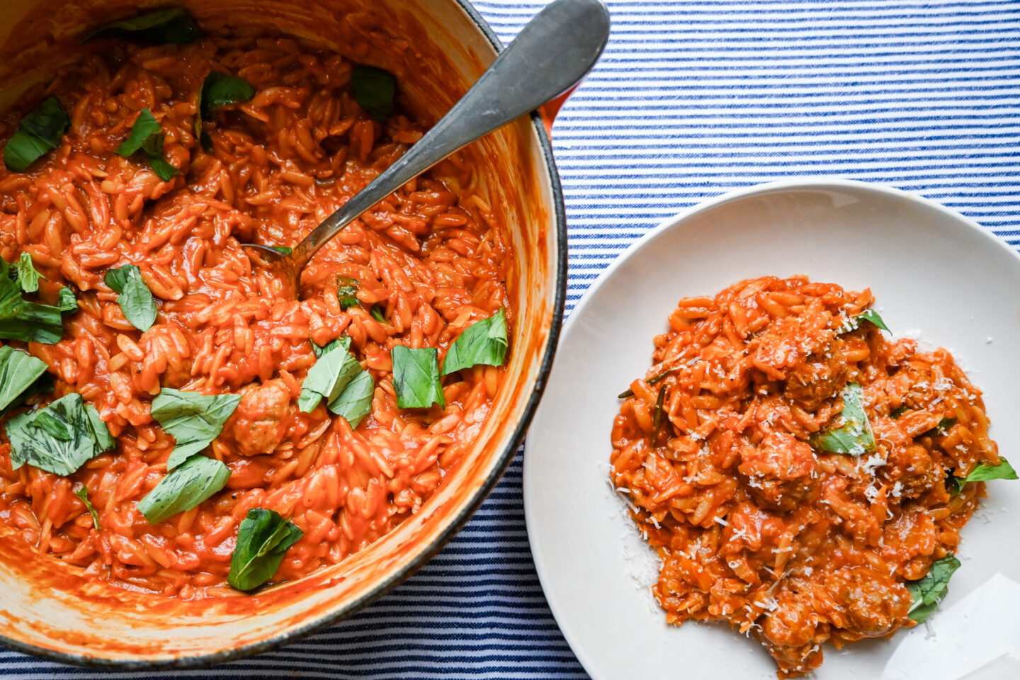 One Pot Sausages Meatballs With Orzo in a tomato sauce sprinkled with torn basil leaves in an orange Le Creuset Casserole next to a pasta bowl with a serving sprinkled with Parmesan Cheese