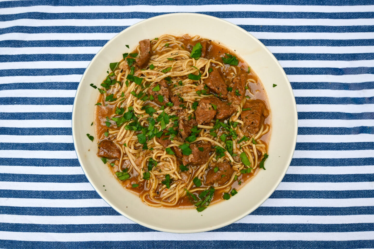 White bowl of beef cubes & ramen egg noodles in a broth sprinkled with parsley