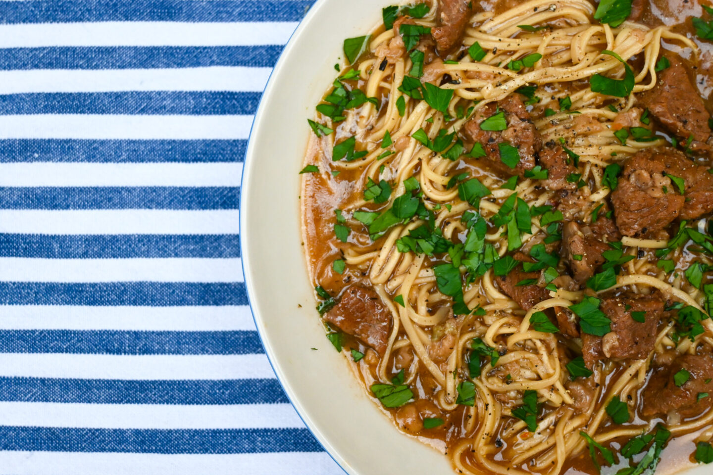 White bowl of beef cubes & ramen egg noodles in a broth sprinkled with parsley