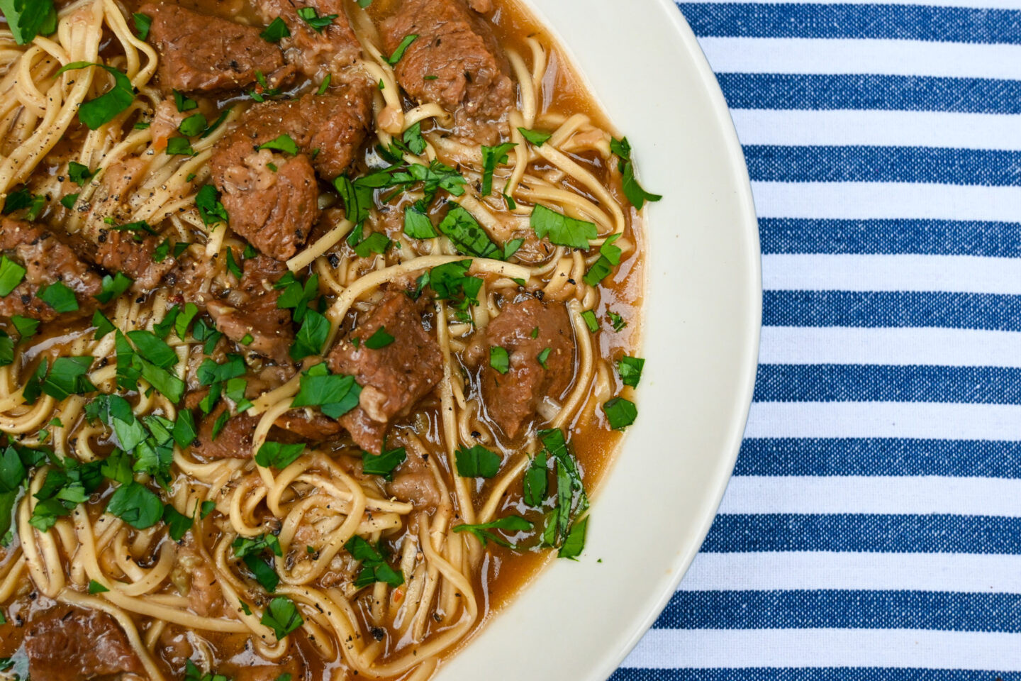 White bowl of beef cubes & ramen egg noodles in a broth sprinkled with parsley